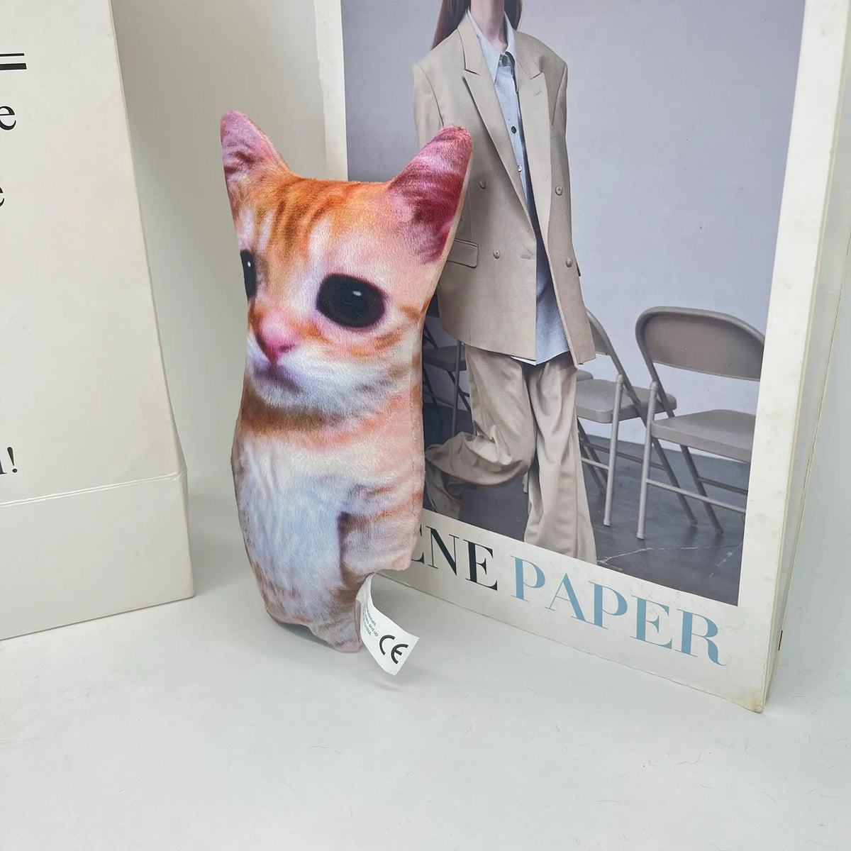 A small orange tabby cat plush doll is displayed in a box next to a photo of a person wearing a beige jacket.