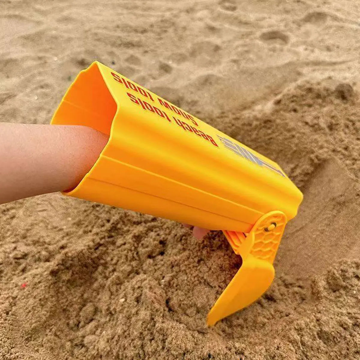 An orange kids excavator digging toy is shown in the palm of a hand, resting on sand.