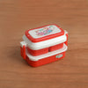 A miniature doll picnic lunch box with a red and white design sits on a wooden surface.