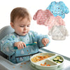 A baby is eating food from a bib while surrounded by various full-sleeve waterproof baby bibs.