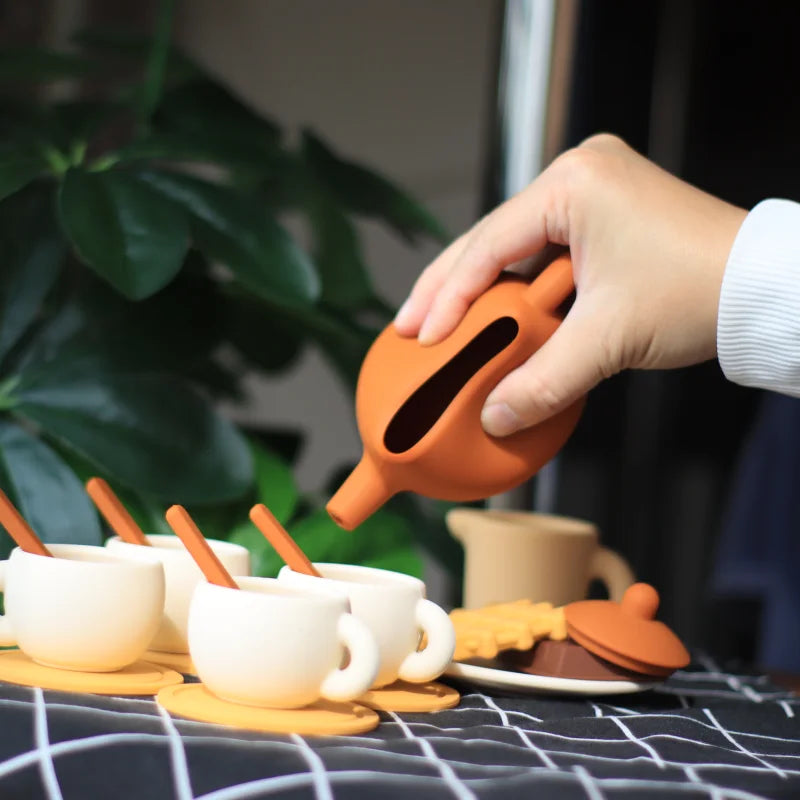 An orange silicone pretend play tea set with a spout is being held over a collection of small cups and saucers.