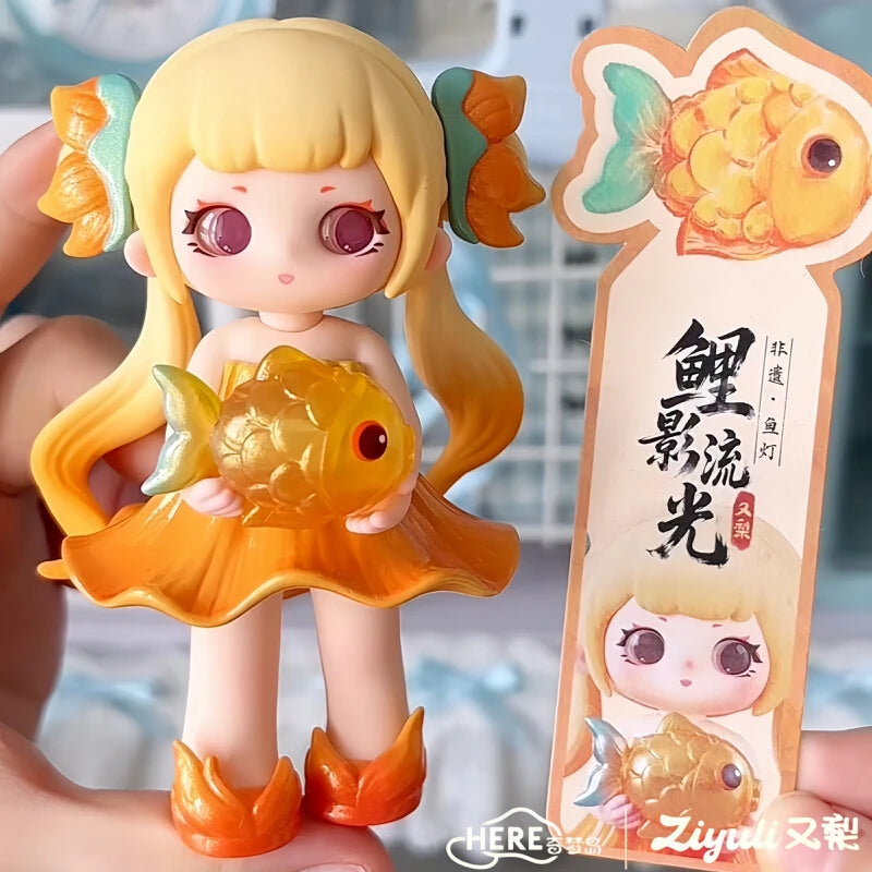A figurine of a girl with blonde hair and large eyes holding a golden fish, packaged in a cardboard box.
