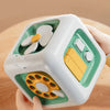 A hand holds a white cube Montessori busy toy with green accents and yellow components.