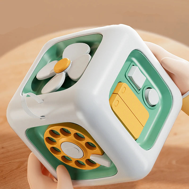A hand holds a white cube Montessori busy toy with green accents and yellow components.