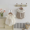 A beige sleeveless cotton jumpsuit for a baby girl with a white lace collar hanging on a wall rack.