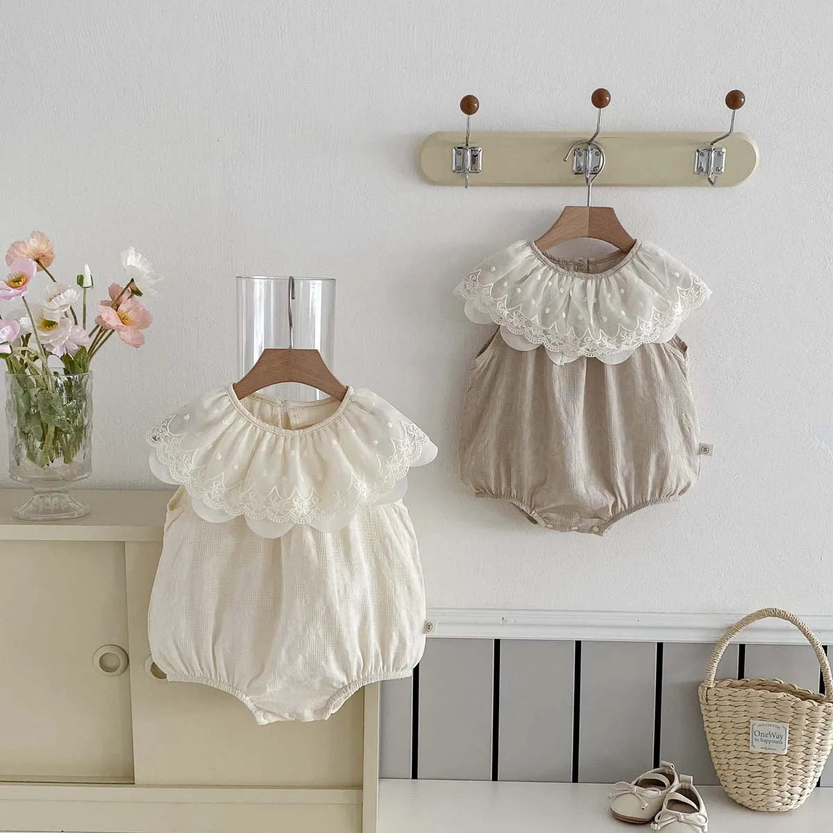 A beige sleeveless cotton jumpsuit for a baby girl with a white lace collar hanging on a wall rack.