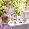 Three young girls play with a wooden pretend tea party set outdoors in a sunny garden setting.