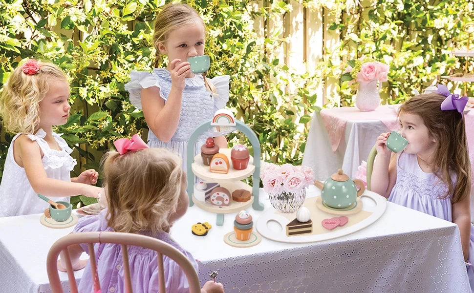 Three young girls play with a wooden pretend tea party set outdoors in a sunny garden setting.