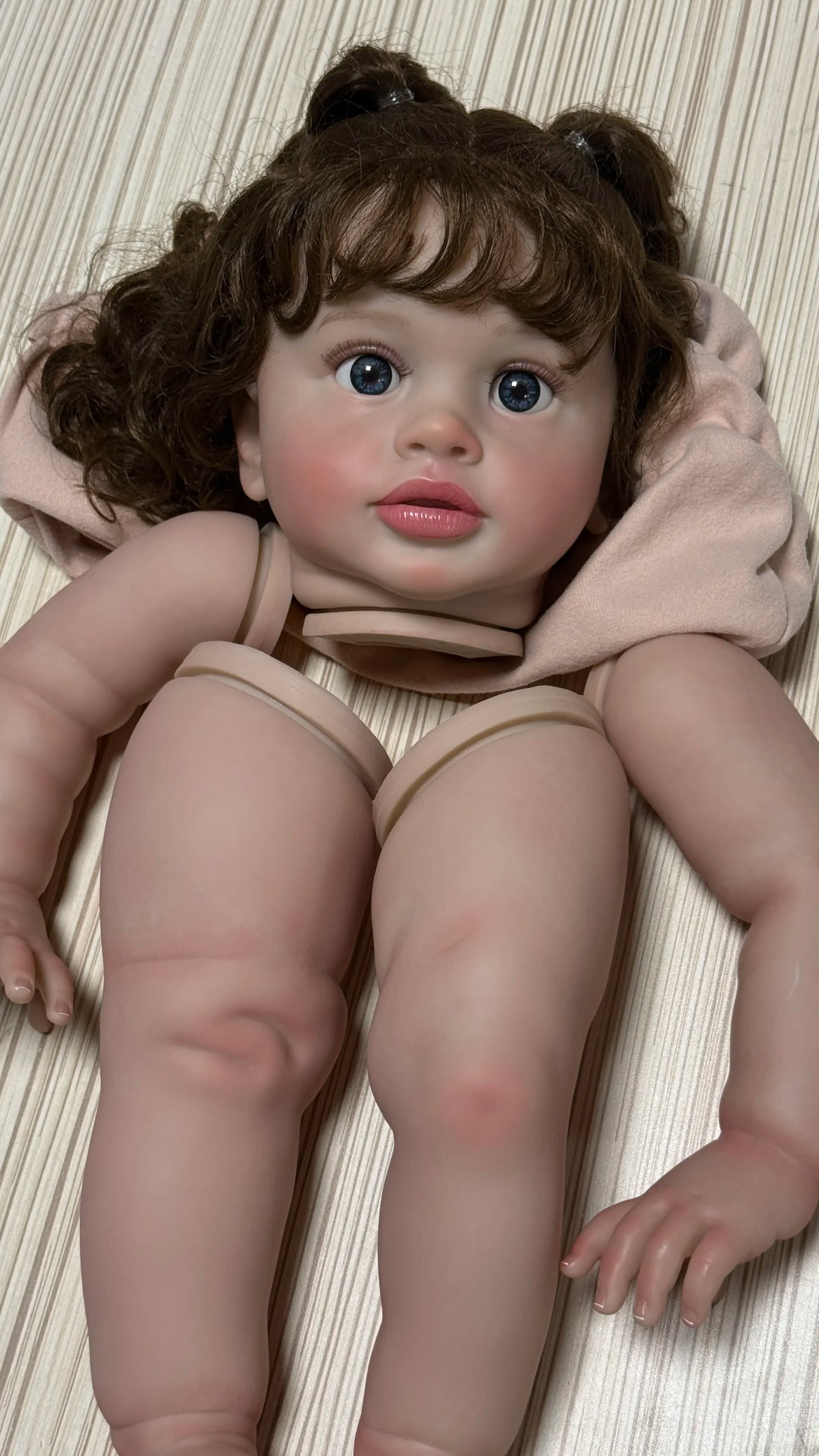 A lifelike reborn baby doll kit featuring a baby with brown hair and blue eyes wearing a pink hooded garment.