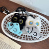 A cute plush black cat keychain charm with blue and white accents displayed in a white basket.