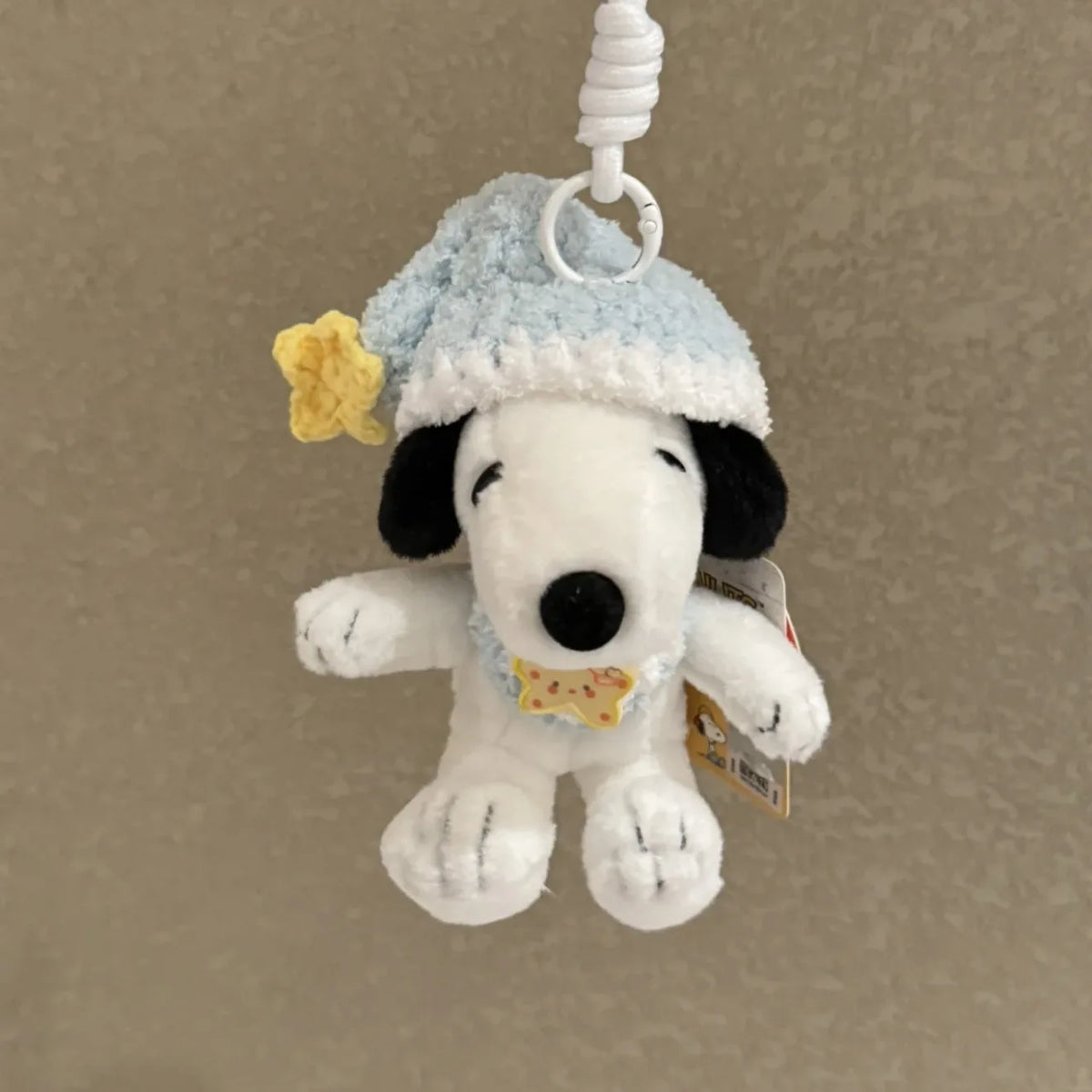 A white dog plush toy wearing a light blue knitted hat and a yellow flower, attached to a white cord for use as a keychain.