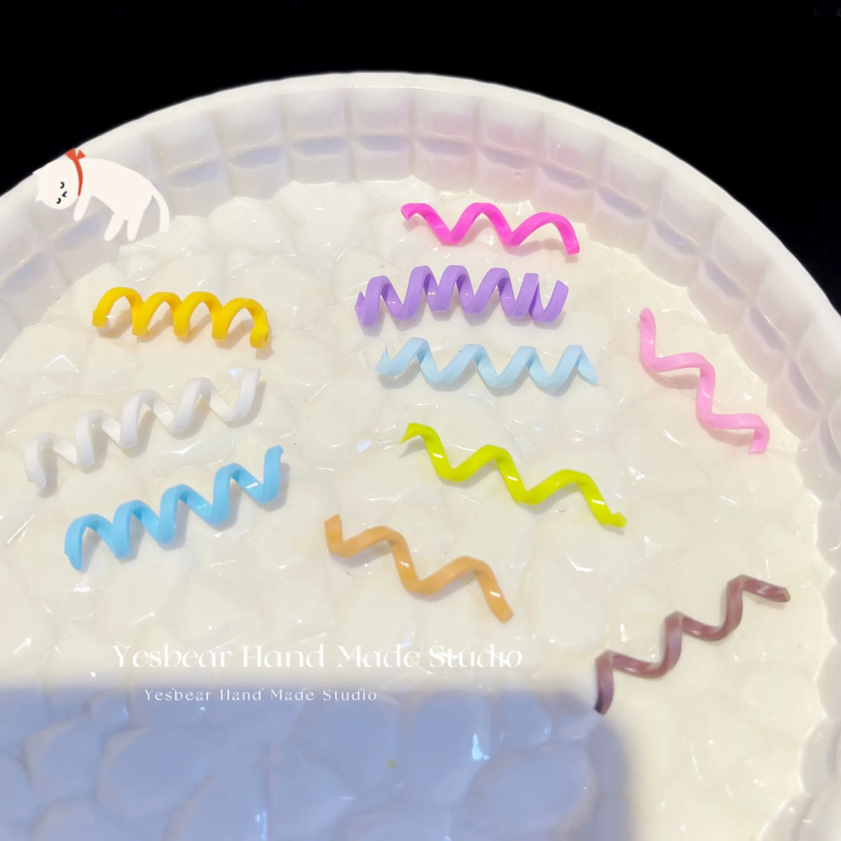 A collection of ten colorful polymer clay marshmallow charms shaped like wavy lines are displayed in a white bowl.