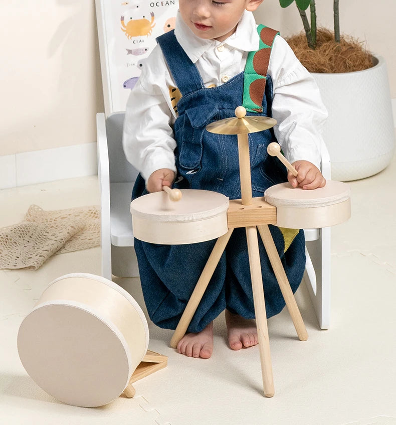 A toddler is sitting in a chair with a wooden drum set, featuring various sized wooden drums and mallets.