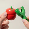 A hand holds a red wooden apple and a green wooden knife, part of a magnetic fruit cutting play set.