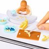 A miniature baby food doll surprise box showing a baby doll, yellow bananas, and a wooden serving tray.