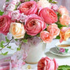 A bouquet of pink and peach roses, sunflowers, and peonies arranged in a vase on a white surface.