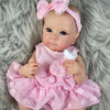 A pink reborn baby doll with painted hair and a bow, wearing a matching pink dress, lying on gray fur.