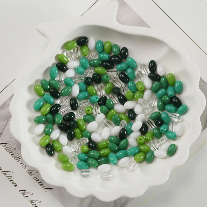 A white oval dish filled with various green and black glass rice grain beads for jewelry making