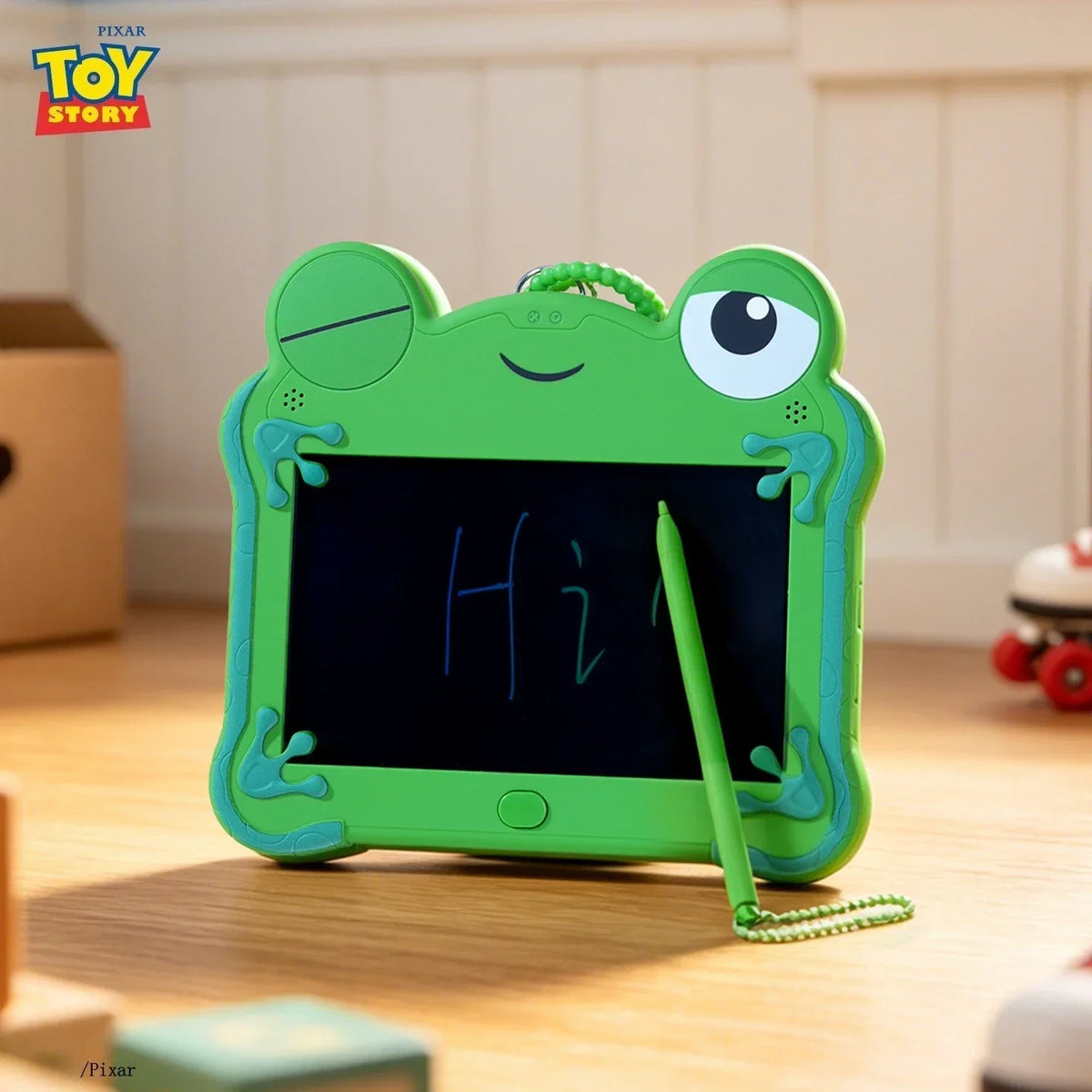 A bright green frog-shaped LCD drawing tablet keychain with a stylus rests on a wooden surface.