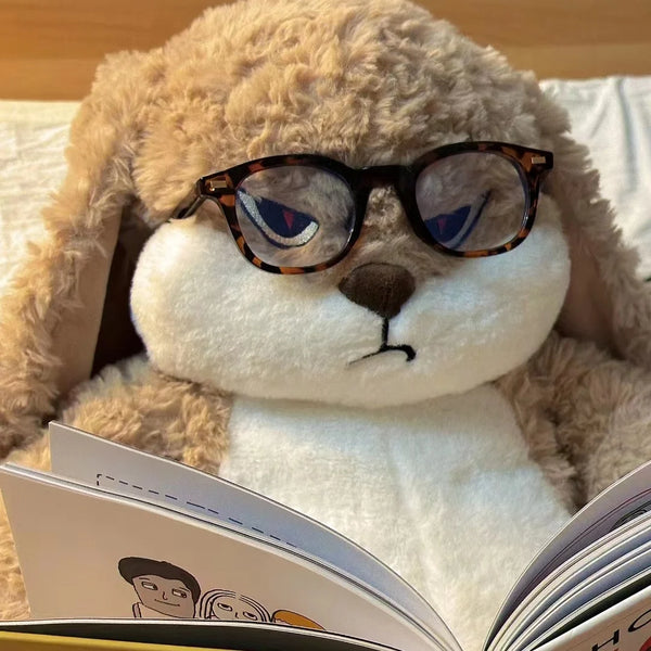 Un grand lapin en peluche doux portant des lunettes rondes regarde un livre ouvert.