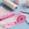 A pink tape measure is laid out on a light blue surface next to a roll of white thread and a pink fuzzy fabric.