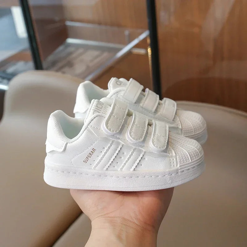 A hand holds a pair of white Adidas Superstar sneakers with hook-and-loop closure.