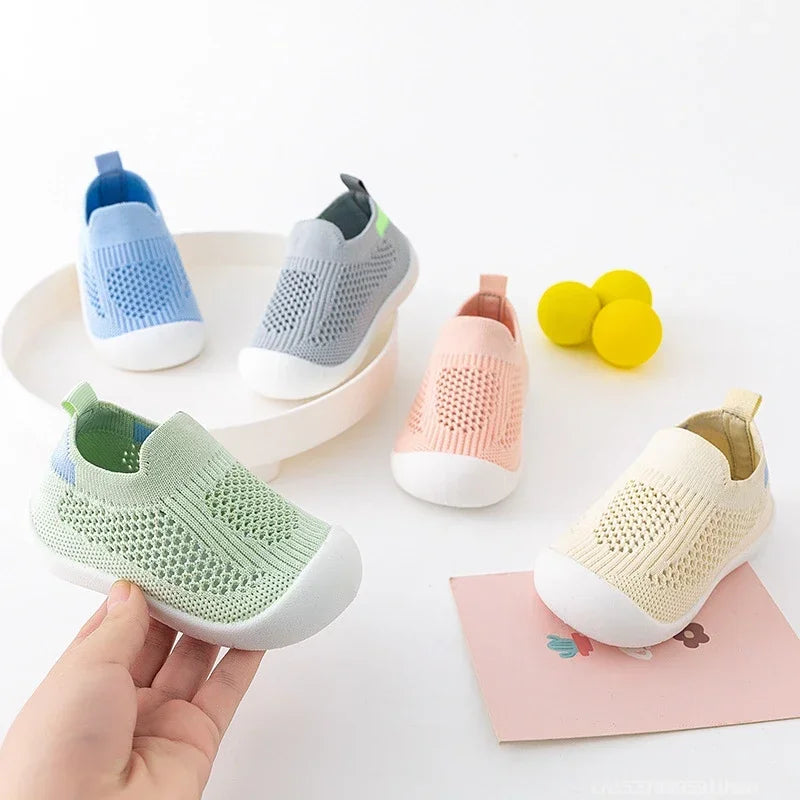A hand holds a pair of breathable mesh slip-on sneakers for toddlers in various pastel colors.
