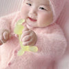 A baby is holding a yellow silicone teether toy while wearing a pink fuzzy hooded garment.