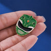 A hand holds a green enamel pin featuring a dinosaur ranger helmet design with gold accents.