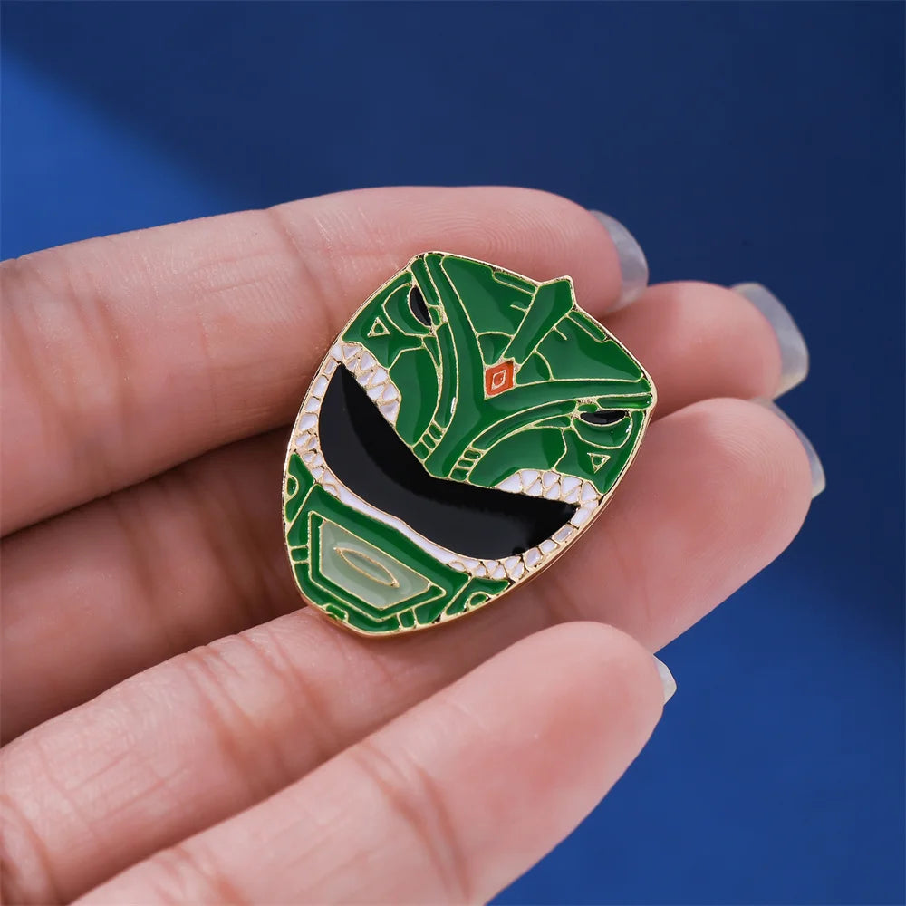 A hand holds a green enamel pin featuring a dinosaur ranger helmet design with gold accents.