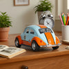 A 1:36 scale die-cast retro Beetle car in light blue and orange sits on a wooden surface next to a potted plant and an alarm 