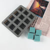 A gray square silicone soap mold with twelve cavities is shown next to several light blue silicone blocks.