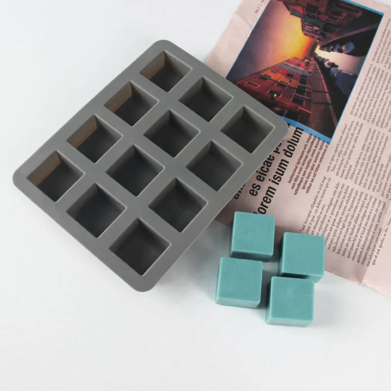 A gray square silicone soap mold with twelve cavities is shown next to several light blue silicone blocks.