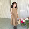 A young girl wearing a sleeveless beige cotton sundress stands in front of a sheer white curtain.