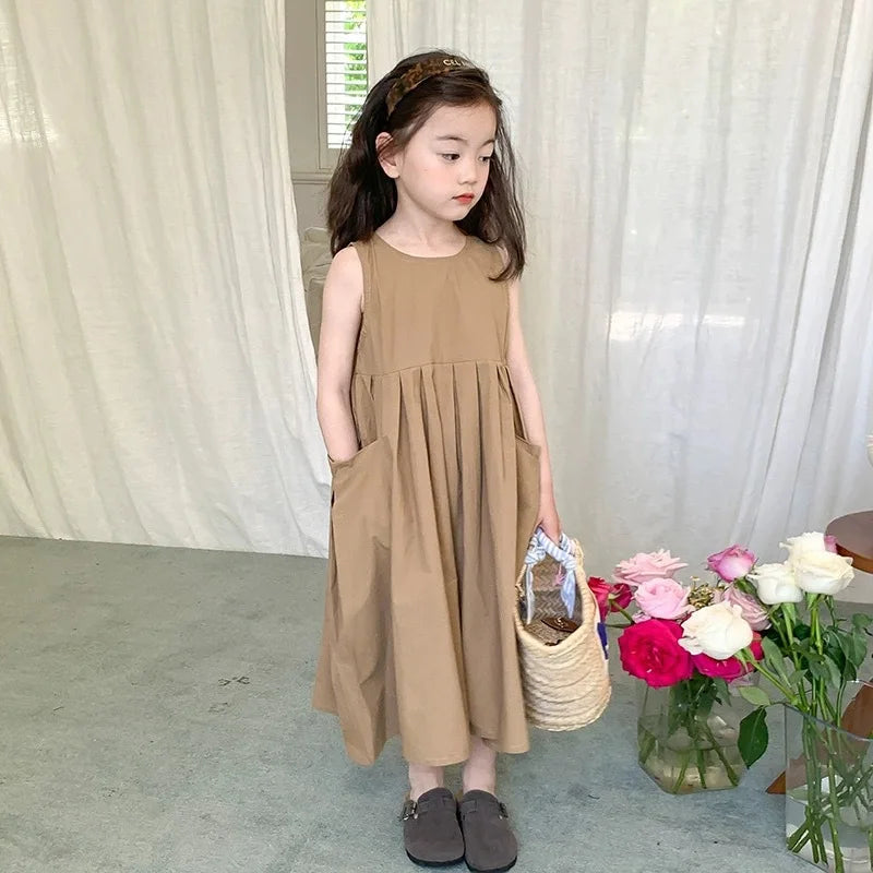 A young girl wearing a sleeveless beige cotton sundress stands in front of a sheer white curtain.