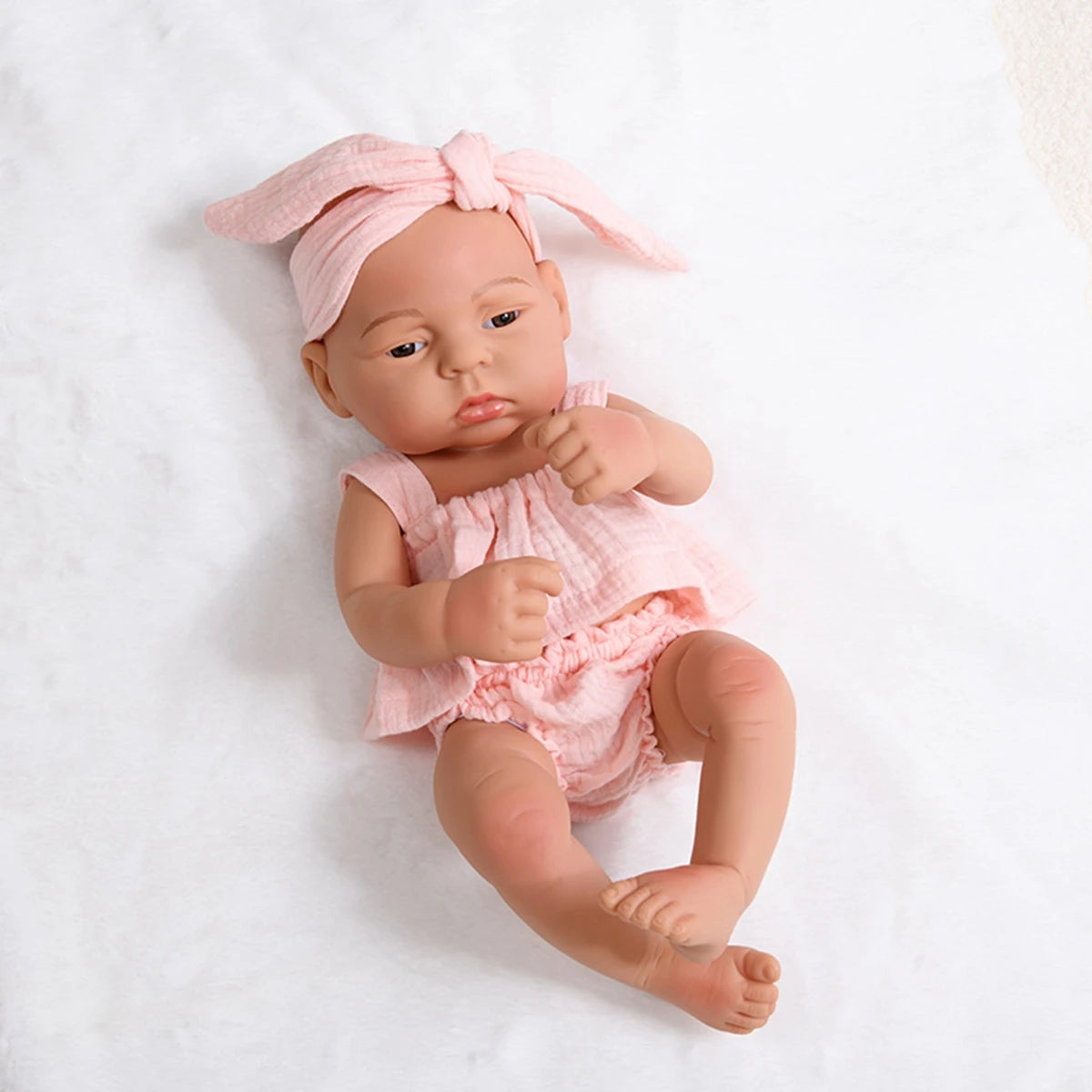 A lifelike vinyl reborn doll with a pink outfit and a matching head wrap, lying on a white surface.