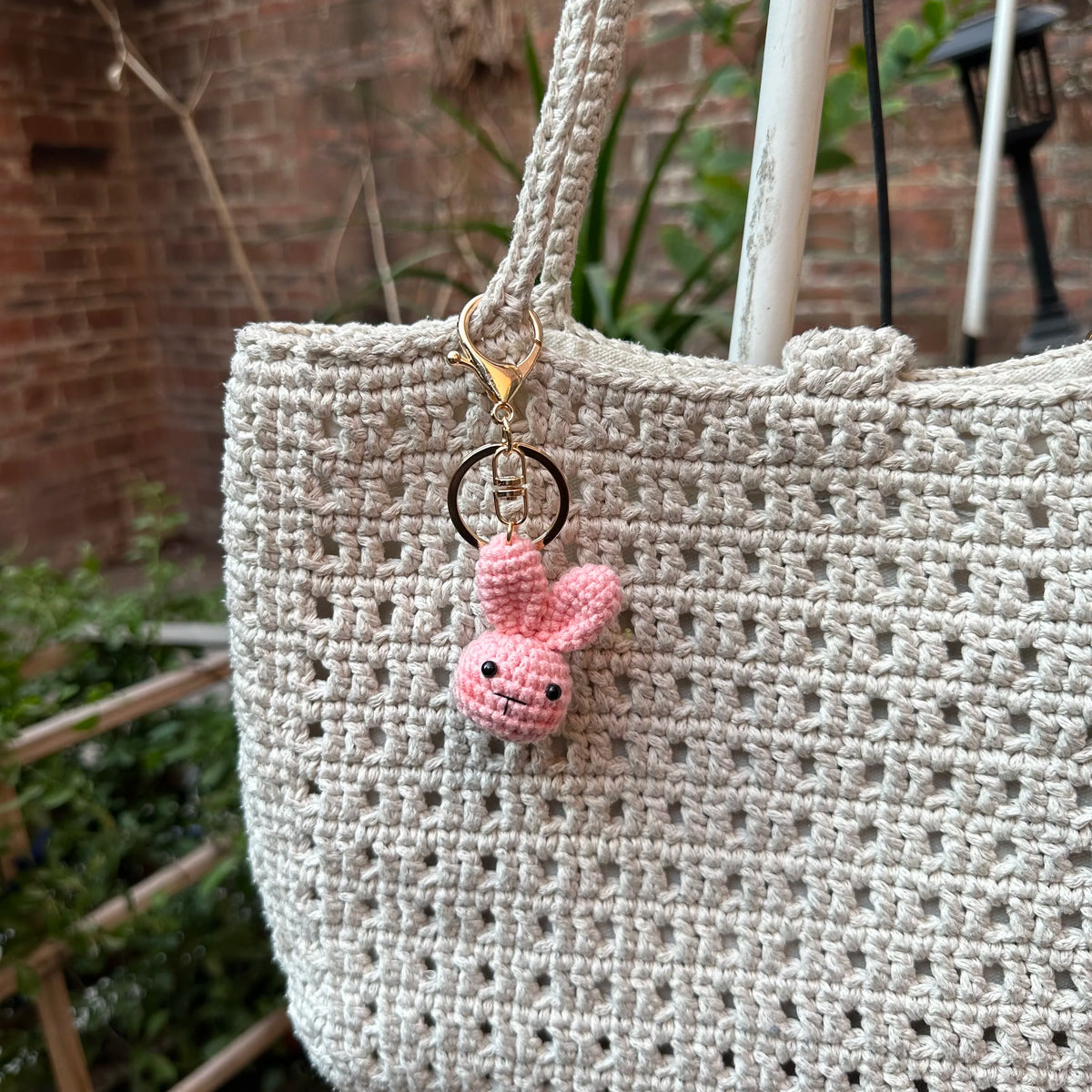 A light beige crocheted bag with a gold-toned keychain featuring a small pink rabbit charm.