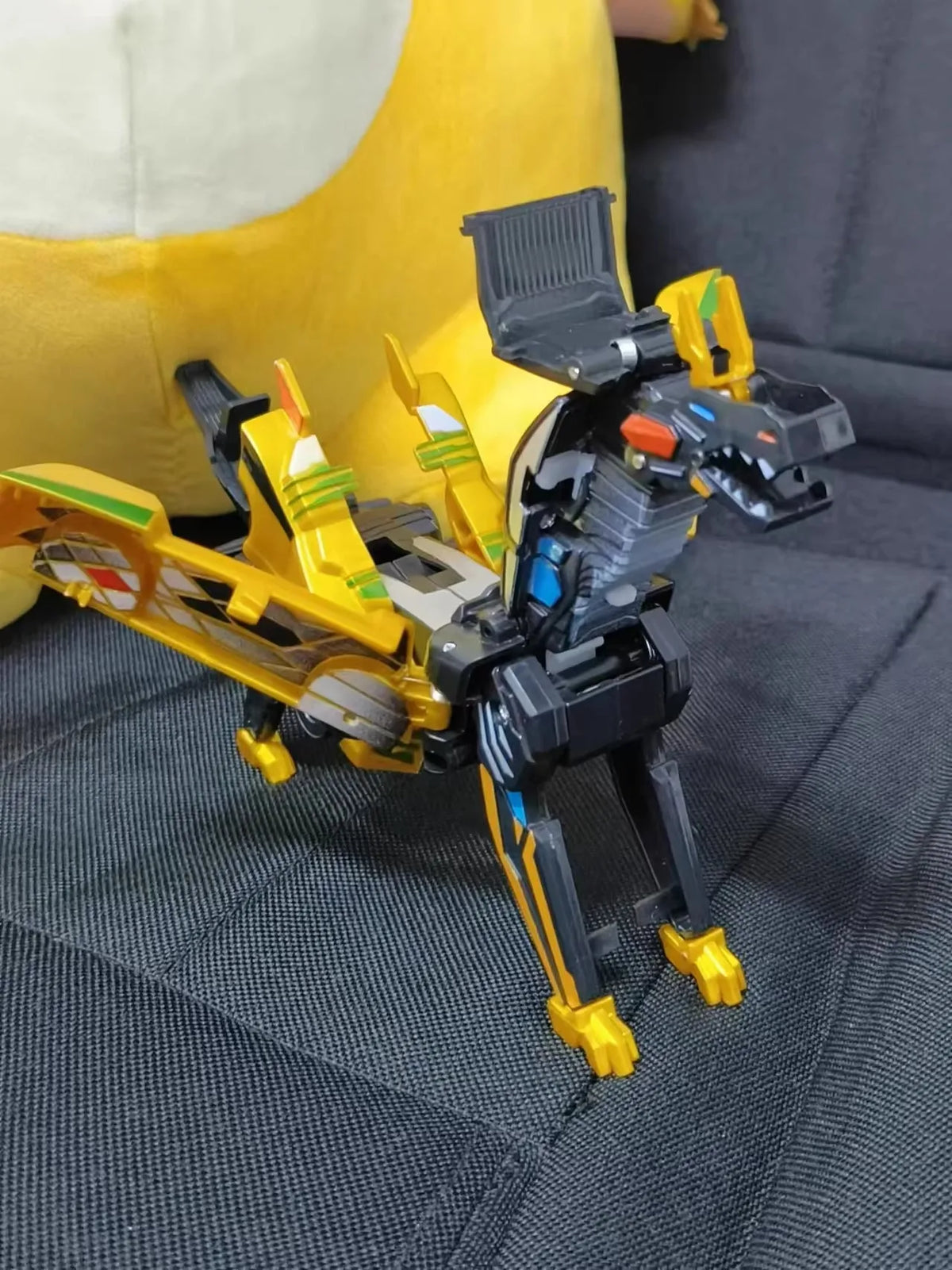 A black and yellow transforming dinosaur car toy is shown resting on a dark gray fabric surface.