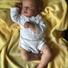 A newborn baby doll with a cloth body is lying on a yellow fabric.