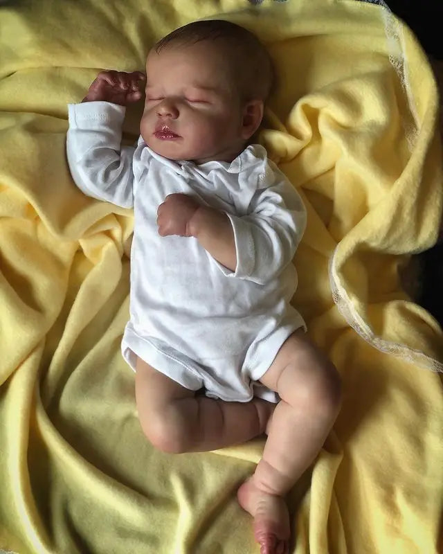 A newborn baby doll with a cloth body is lying on a yellow fabric.