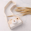 A light wood camera teether toy with a beige strap rests on a marble surface next to wheat stalks.