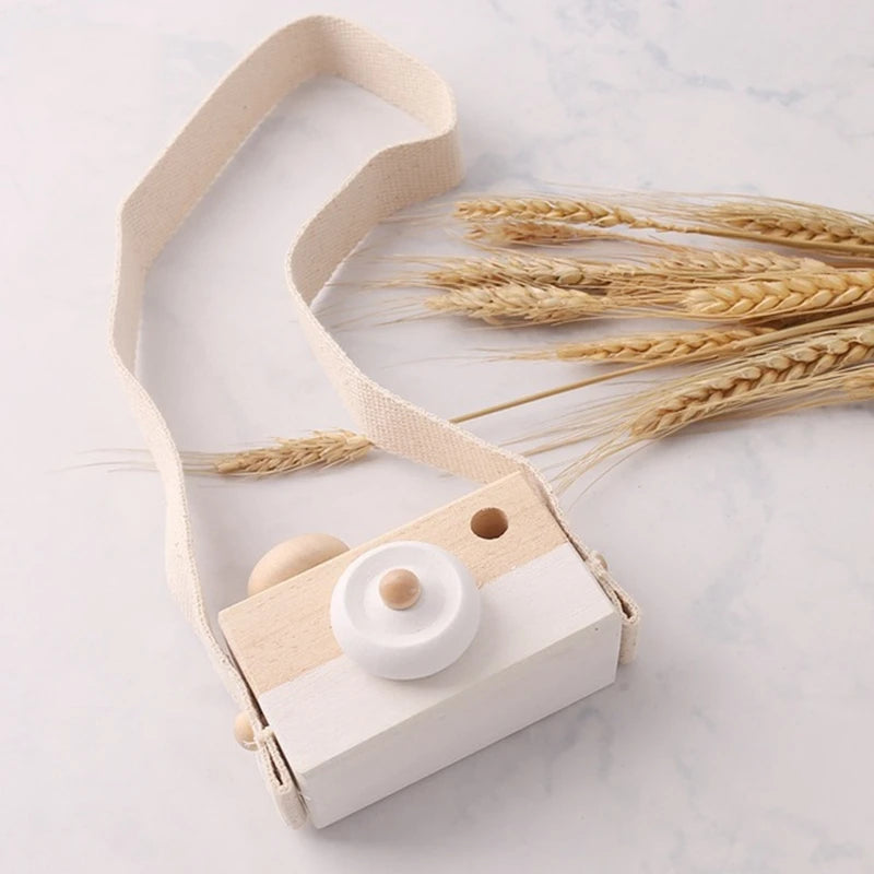 A light wood camera teether toy with a beige strap rests on a marble surface next to wheat stalks.