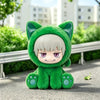 A plush collectible figure of a green cat-like character with white hair sitting on a gray outdoor surface.