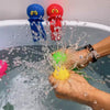 Hands are squeezing a yellow bath toy while water splashes around it in a bathtub with two bath toys visible.