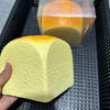 A hand holds a yellow squishy toy shaped like a loaf of bread, next to a clear plastic container.