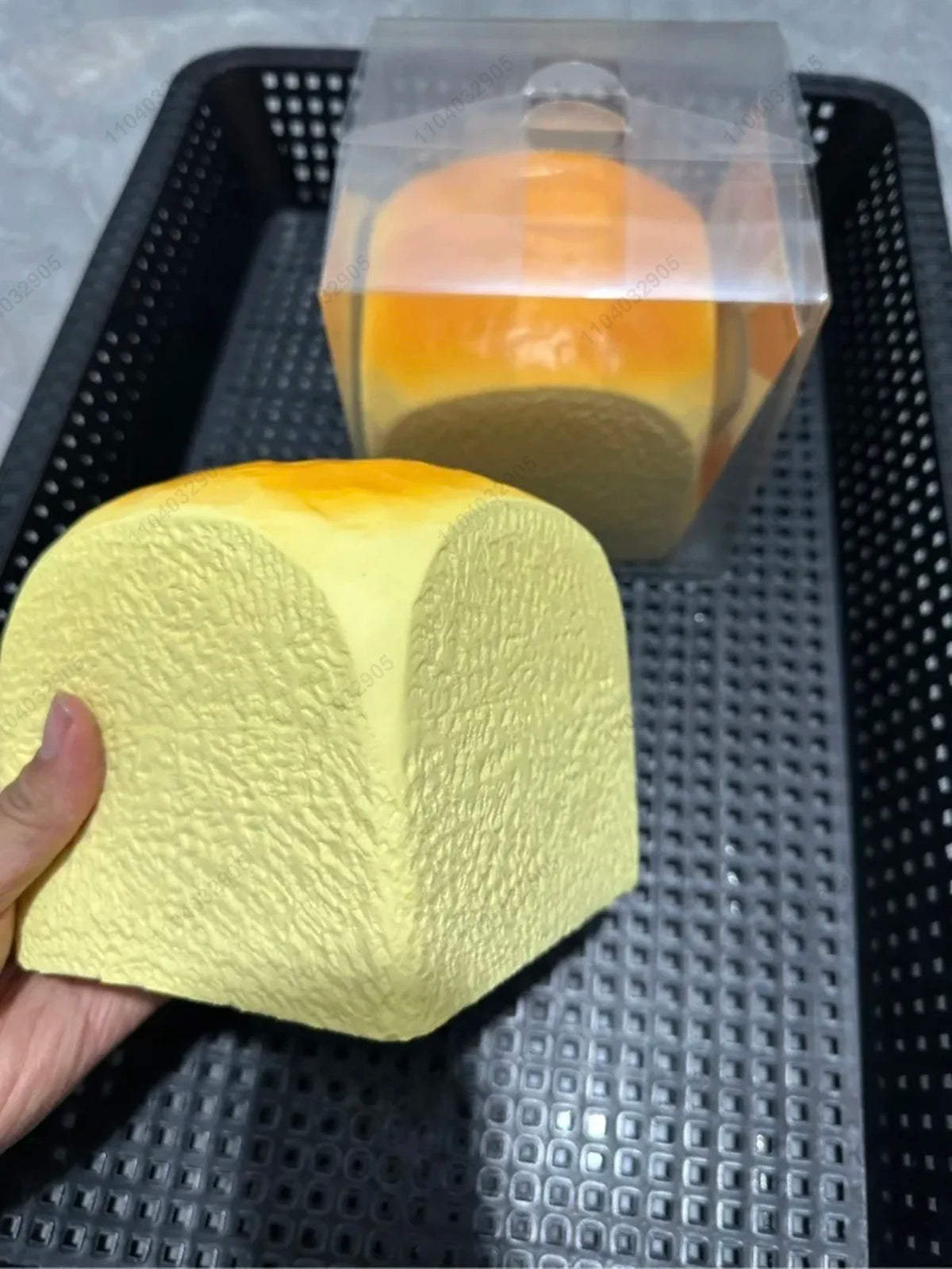 A hand holds a yellow squishy toy shaped like a loaf of bread, next to a clear plastic container.