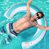 A man wearing sunglasses is floating on a striped U-shaped inflatable pool float in clear blue water.