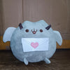 A light blue plush cat doll with horns and a pink heart-shaped envelope on its chest.