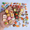 A hand holds a collection of miniature food items including cakes, wine, and various snacks on a white background.