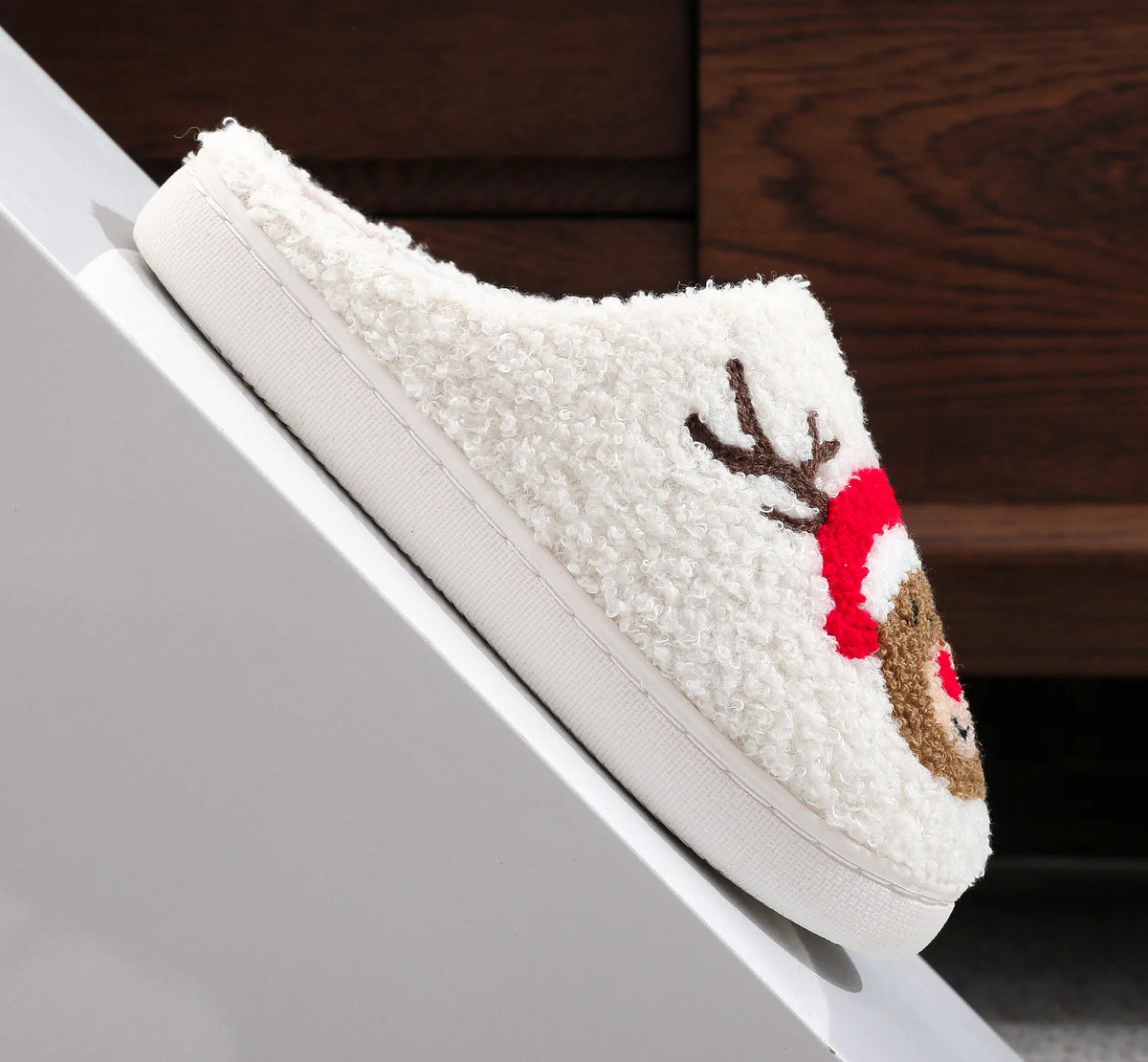 A white plush non-slip indoor slipper with a reindeer design is shown resting on a white surface.
